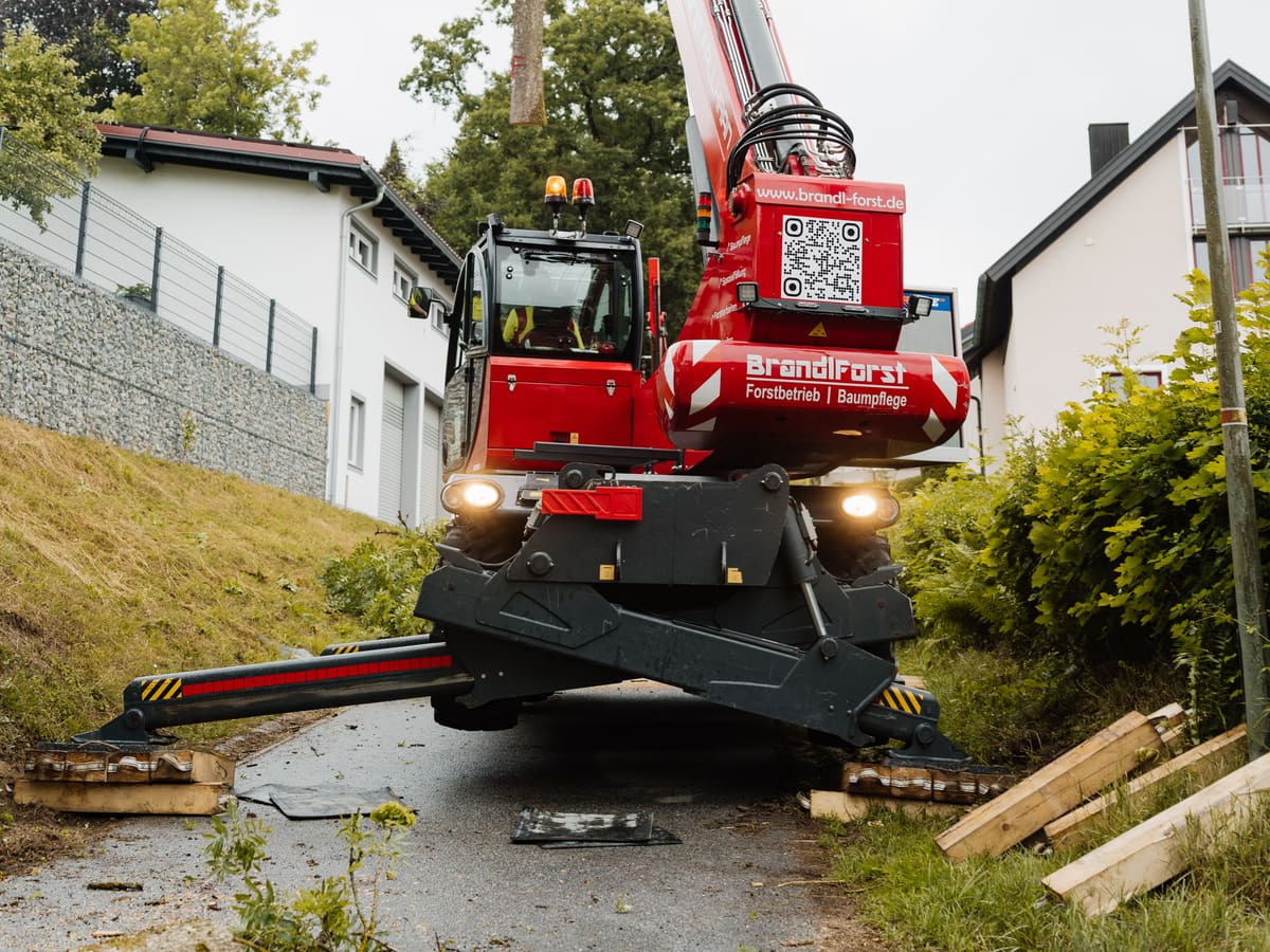 Magni RTH 6.30 Fällkran voll abgestützt zwischen zwei Wohnhäusern, Ausleger zum Baum ausgefahren